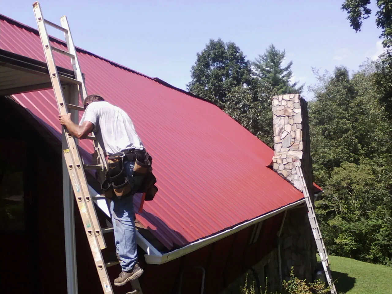 Roofer on a ladder beside a red metal roof and stone chimney — Soffit & Fascia Repair by Roofing Brothers in Tobaccoville